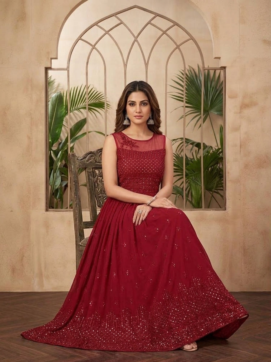 A woman models a sleeveless deep red evening gown with a sheer embroidered bodice and a full flared skirt, posed in front of a beige arched wall with decorative plants celebrating Diwali.
