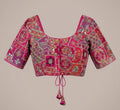 A vibrant magenta pink and purple embroidered blouse with intricate gold and silver mirror work, displayed against a solid beige background.