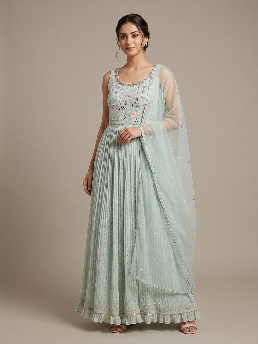 A woman models a light mint green anarkali gown with a matching sheer dupatta. The sleeveless dress features a heavily embroidered bodice with floral details in peach, pink, and green, and a pleated skirt with a ruffled hem.